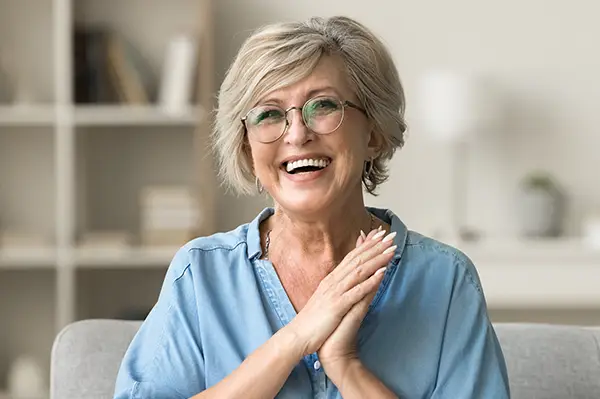 Cheerful pretty older woman in elegant glasses sitting on cozy home couch with dental implants from D.A. Dental in Auburn, MA