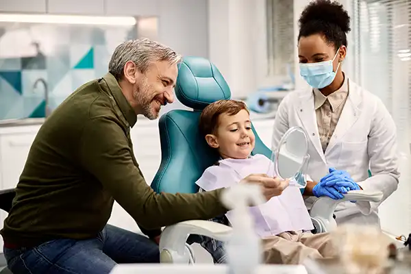 Father holding a mirror for his happy, young son sitting in a dental chair at D.A. Dental in Auburn, MA