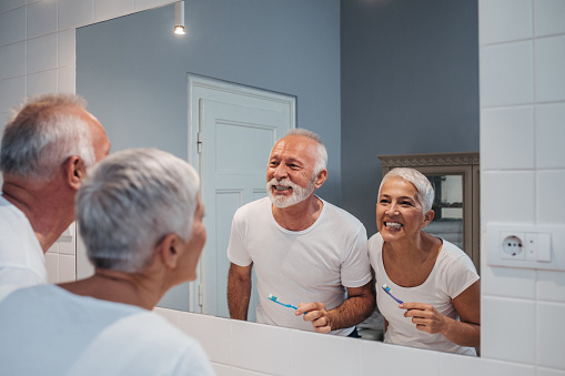 Elderly couple smiling and brushing their teeth in the bathroom mirror at D.A. Dental in Auburn, MA